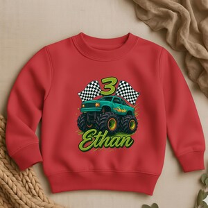 May include: A red sweatshirt featuring a green monster truck design with the number 3, checkered flags, and the name "Ethan" in a green, stylized font. The monster truck has large black tires with yellow rims.