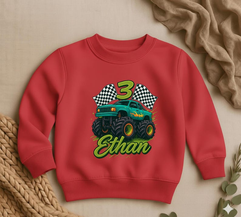 May include: A red sweatshirt featuring a green monster truck design with the number 3, checkered flags, and the name "Ethan" in a green, stylized font. The monster truck has large black tires with yellow rims.