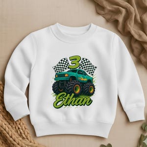 May include: White sweatshirt featuring a green monster truck design with the number 3, checkered flags, and the name "Ethan" in green script. The monster truck has large black tires with yellow rims.