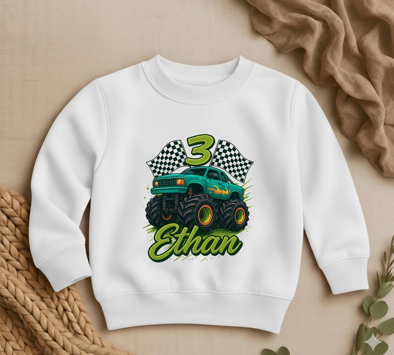 May include: White sweatshirt featuring a green monster truck design with the number 3, checkered flags, and the name "Ethan" in green script. The monster truck has large black tires with yellow rims.