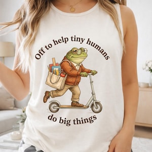 May include: A white sleeveless shirt featuring a graphic of a frog wearing glasses and a brown cardigan, riding a scooter while carrying a tote bag filled with school supplies. The text reads, Off to help tiny humans do big things.