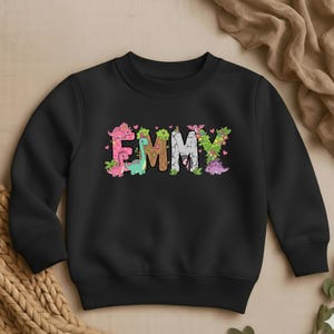 May include: Black crewneck sweatshirt with the name "EMMY" in colorful, dinosaur-themed lettering. The letters are decorated with pink, green, and brown dinosaurs, flowers, and leaves. A cute and playful design for kids.