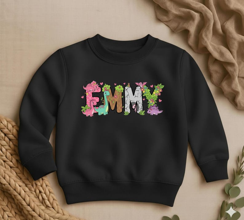 May include: Black crewneck sweatshirt with the name "EMMY" in colorful, dinosaur-themed lettering. The letters are decorated with pink, green, and brown dinosaurs, flowers, and leaves. A cute and playful design for kids.