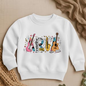 May include: White crewneck sweatshirt featuring the word "ARIA" in colorful musical instrument designs. The design includes a pink guitar, piano keys, a gold saxophone, and a brown acoustic guitar. The word "SING" is also included.