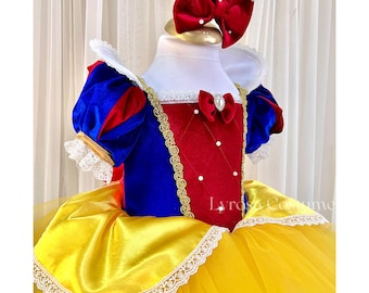 May include: A Snow White-inspired costume featuring a blue and red bodice with gold trim, a yellow skirt, and a red bow headband. The costume is designed for children and includes a white collar and lace details.