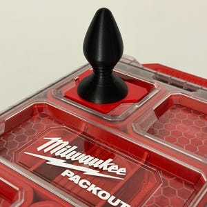 Op de afbeelding: Een zwart, druppelvormig object met een uitlopende basis, rustend op een rood vierkant. Het object staat bovenop een rode en doorzichtige plastic opbergdoos. De doos heeft de witte tekst "Milwaukee PACKOUT".