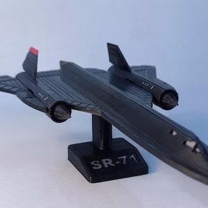 Puede incluir: Maqueta negra del avión SR-71 Blackbird, impresa en 3D y montada sobre un soporte. El modelo presenta detalles como alas, aletas de cola y góndolas de motor. El soporte está etiquetado como "SR-71".