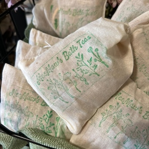 May include: Close-up of several off-white linen bags filled with bath tea. Each bag has a green printed design of botanical illustrations and the text "MeeMaw's Bath Teas". The bags are in a black wire basket.