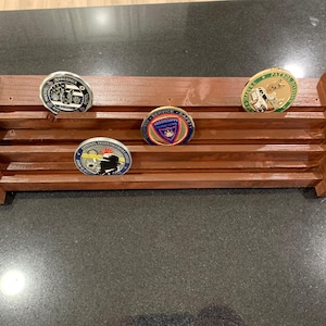 Op de afbeelding: Een bruine houten display plank met vier challenge coins. De plank heeft drie horizontale sleuven. De munten hebben verschillende ontwerpen en kleuren, waaronder blauw, goud en zilver. De plank is ontworpen om verzamelmunten te bewaren en te presenteren.