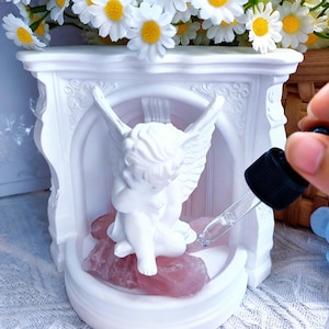 Könnte beinhalten: Eine weiße Cherub-Statue sitzt auf rosa Rosenquarzsteinen in einer dekorativen weißen Nische. Eine klare Pipette wird über den Steinen gehalten. Weiße Gänseblümchen mit gelben Zentren befinden sich im Hintergrund.