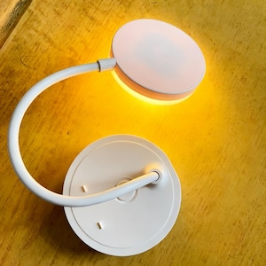 Puede incluir: Lámpara de escritorio LED blanca con cabezal y base circulares. La lámpara tiene un brazo flexible y curvado y emite una luz amarilla cálida. La lámpara está diseñada para uso en escritorio.