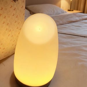 Puede incluir: Una lámpara ovalada y brillante que emite una luz suave y cálida. La lámpara tiene una parte superior redondeada y una base oscura. La luz se difunde, creando un ambiente suave, adecuado para una mesita de noche o iluminación ambiental.