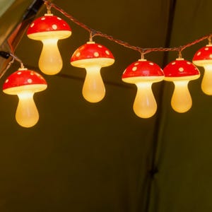 Puede incluir: Una cadena de luces decorativas con forma de setas rojas con lunares blancos. Los sombreros de las setas son rojos y los tallos son de un amarillo cálido y brillante. Las luces están ensartadas en un fino cable marrón.