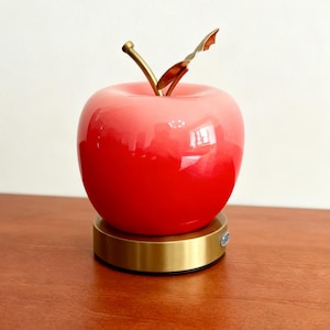 Può includere: Un oggetto decorativo a forma di mela rossa e rosa, con uno stelo e una foglia dorati. La mela poggia su una base circolare dorata. Potrebbe essere una lampada o un oggetto decorativo.