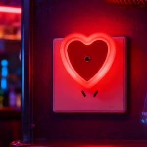 Può includere: Una luce notturna a forma di cuore rosso collegata a una presa elettrica bianca. La luce notturna ha un contorno rosso brillante e un centro rosso pieno. La presa è montata su una superficie scura.