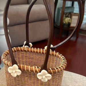 May include: A woven basket bag with a light tan natural reed body and a dark brown rim. It features two dark brown leather handles attached to the basket with four white, four-petaled flower-shaped accents at the base of the handle straps.