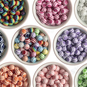 K&ouml;nnte beinhalten: Zw&ouml;lf wei&szlig;e Schalen gef&uuml;llt mit bunten, gestreiften Perlen. Jede Schale enth&auml;lt eine andere Farbkombination von Perlen, darunter Regenbogen, rosa und wei&szlig;, blau und wei&szlig;, rot und wei&szlig;, gr&uuml;n und wei&szlig;, lila und wei&szlig;, orange und wei&szlig;, gelb, schwarz und wei&szlig; und wei&szlig;.