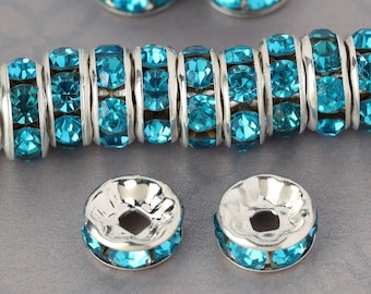 May include: A close-up of turquoise blue rhinestone bead spacers. The spacers are silver-toned and have a round shape. There are several spacers scattered on a gray surface, and a strand of the spacers is laid out horizontally.