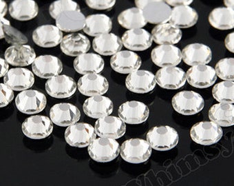 May include: A collection of small, clear, round rhinestones scattered on a black surface. The rhinestones are faceted, reflecting light, and appear to be made of glass or crystal. The image has the text "Whimsy & Pop" in the corners.