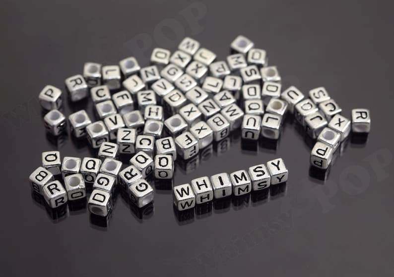 May include: A close-up of silver letter beads spelling out the word "WHIMSY". The beads are scattered on a black surface.