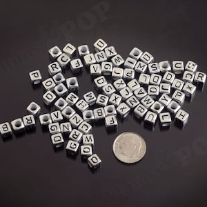 May include: A pile of silver metal letter beads, each bead is a cube with a letter of the alphabet printed on each side. The beads are scattered on a black surface.
