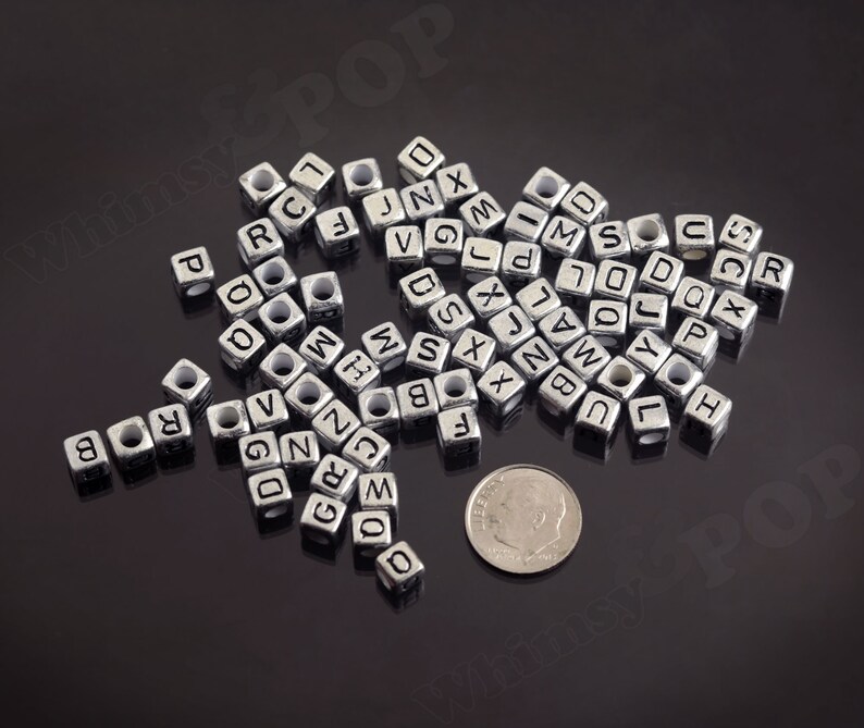 May include: A pile of silver metal letter beads, each bead is a cube with a letter of the alphabet printed on each side. The beads are scattered on a black surface.