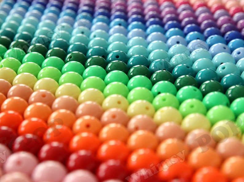 May include: A rainbow assortment of round, glossy plastic beads. The beads are arranged in rows, with each row featuring a different color. The colors range from purple to pink, orange, yellow, green, blue, and turquoise.