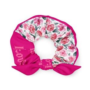 May include: A bright pink scrunchie with a floral pattern and the word "Love" printed on the side. The floral pattern features pink roses and green leaves on a white background. The scrunchie is a hair accessory.