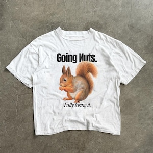 Puede incluir: Una camiseta blanca de manga corta con un gráfico de una ardilla marrón sosteniendo una nuez. El texto "Going Nuts." está impreso en negro sobre la ardilla, y el texto "Fully losing it." está impreso en negro debajo de la ardilla.