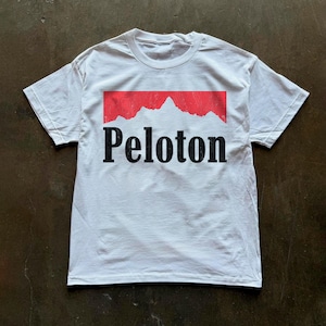 May include: A white short-sleeve t-shirt featuring a graphic on the chest. The graphic consists of a red rectangular shape with a jagged mountain silhouette at the top and the word Peloton printed in black serif font underneath the mountain.