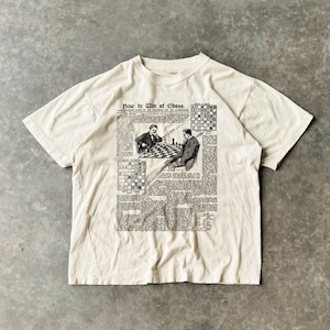 以下が含まれることがあります： クリーム色のTシャツ。白黒の新聞風グラフィックで、「Now to Win at Chess」の文字とチェスの図が描かれています。チェスをする2人の男性が描かれています。