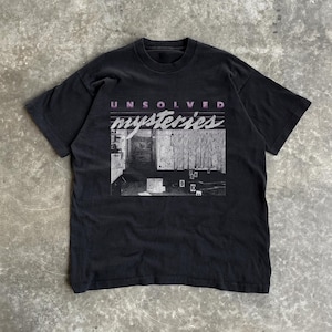 Puede incluir: Camiseta negra con el texto "UNSOLVED mysteries" en rosa y blanco. El gráfico incluye una imagen en blanco y negro de una escena del crimen con marcadores de evidencia. La camiseta es de estilo vintage.