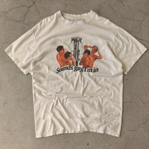 Peut inclure: Un t-shirt à manches courtes de couleur crème avec une impression graphique représentant quatre personnes sous une douche. Sous le graphique, le texte "Sounds gay, I'm in" est écrit en caractères gras noirs sans empattement. Col rond classique.