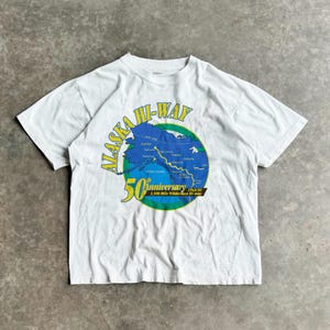 May include: White t-shirt featuring a map of Alaska in blue and green, with the text "ALASKA HI-WAY" in green and yellow. The shirt commemorates the 50th Anniversary of the 1,500 Mile Wilderness Hi-way, 1942-92.