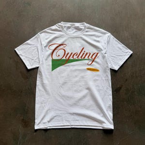 May include: White t-shirt with the word "Cycling" in a cursive, reddish-brown font. A green triangle with a white outline is behind the word. Below the word is a small, yellow oval with text. The shirt is on a gray surface.