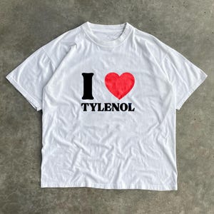 May include: White t-shirt with the text "I ♥ TYLENOL" in black and red. The "I" is black, the heart is red, and the word "TYLENOL" is in a bold, black font. The shirt has a crew neck and short sleeves.