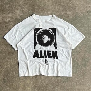 Puede incluir: Camiseta blanca con un gráfico en blanco y negro de una persona con un casco espacial, con la palabra "ALIEN" impresa debajo. Una pequeña figura alienígena está debajo de la palabra. La camiseta es de material suave.