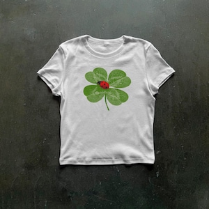 May include: A white short-sleeved t-shirt featuring a central graphic of a green four-leaf clover with water droplets on its surface. A small red ladybird with black spots is positioned on the centre of the clover leaves.