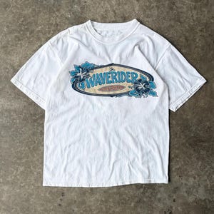 May include: A white short-sleeve t-shirt featuring a central graphic of a surfboard shape with the text WAVERIDER and HAWAII. The design includes blue hibiscus flowers on the left and right sides of the oval frame with a tan background.