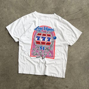 May include: A white short-sleeve t-shirt featuring a graphic of a Las Vegas slot machine with the text "Las Vegas" and "$1". The slot machine display shows three sevens in blue and the word "BAR" in red, with silver coins scattered below.