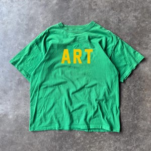 Peut inclure: Un t-shirt vert avec le mot "ART" imprimé en lettres jaunes sur la poitrine. Le t-shirt est fait d'une matière douce et a des manches courtes et un col rond. Le t-shirt est présenté sur une surface grise.