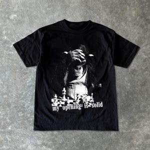 Può includere: T-shirt nero con grafica usurata di uno scimpanzé che si copre gli occhi, una scacchiera e la scritta "my opening is solid" in bianco. La maglietta è realizzata in materiale morbido.