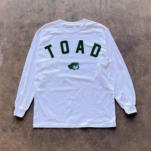 May include: White long-sleeve shirt with the word "TOAD" in green block letters across the chest. Below the word "TOAD" is a green graphic of a toad. The shirt has a crew neck and long sleeves.