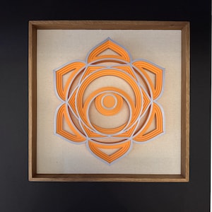 May include: Orange and silver layered geometric art piece in a wooden frame. The artwork features a symmetrical design with a central circular element surrounded by petal-like shapes. The piece is mounted on a beige background.