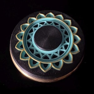 May include: A round, layered decorative object with a floral design. The base is black with a gold edge. The top layer features a teal and gold floral pattern, with a central teal circle and a black center.
