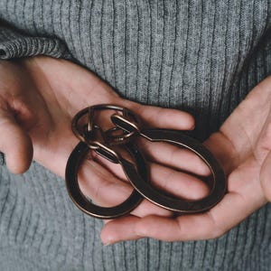 May include: Three bronze-coloured metal keychains, each with an oval ring and a connecting clasp. The keychains are held in open hands against a grey ribbed sweater background.
