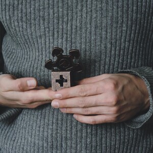 May include: A dark brown metal fidget spinner with a cross design on a silver cube. The spinner is held in front of a grey ribbed jumper.