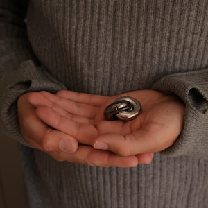 May include: A small, polished, metallic puzzle object consisting of two interlocking, rounded, silver-toned rings. The surface of the metal is smooth and reflective, catching the light to highlight its curved, circular shape and solid construction.