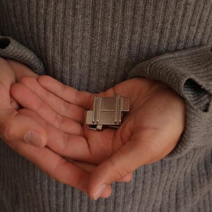 May include: A small, metallic, rectangular puzzle piece with a smooth, silver-toned finish. The object features a series of parallel vertical grooves and a stepped, interlocking design, appearing as a compact, solid, and geometric mechanical component.