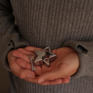 May include: A silver-toned metal padlock shaped like a five-pointed star, featuring a matching small key with a star-shaped handle. The padlock has a central keyhole and a smooth, reflective finish, presented as a set of two distinct metal components.